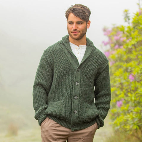 A man stands outdoors in a lush, misty environment, wearing a green knit cardigan over a white shirt. He has short brown hair and is looking slightly to the side, with purple flowers and greenery visible in the background.