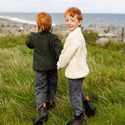 Two young boys stand together in a grassy field. One boy wears a cream-colored kids Aran sweater and looks at the camera, while the other is dressed in a dark green sweater and is running toward the sea. Rocky terrain and a distant ocean in the background.