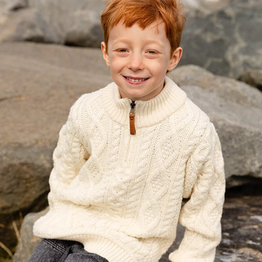 A smiling boy with red hair sits on a rock, wearing a cream-colored kids merino wool Aran sweater. The background features grey rocks, suggesting an outdoor setting.