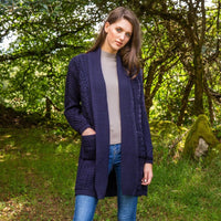 A woman stands in a wooded area, wearing a long navy aran cardigan over a grey top and blue jeans. She has long brown hair and looks towards the camera, with trees and greenery in the background.