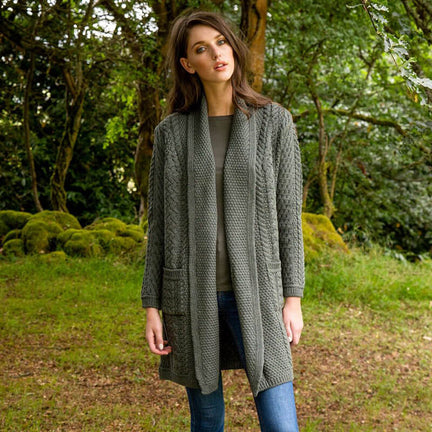 A woman stands outdoors in a green setting, wearing a long, green knitted aran cardigan over a gray top and blue jeans. She gazes at the camera with a neutral expression.