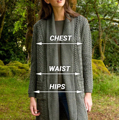 A woman stands in an outdoor setting wearing a long, knitted green aran cardigan over a dark shirt and jeans. Lines labeled 