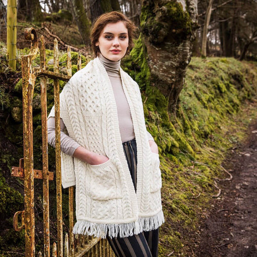 Cream merino wool pocket shawl with cable knit patterns and fringe, worn outdoors beside an old metal gate on a mossy woodland path.