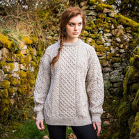 A woman standing on a rocky coast wearing an  oatmeal Womens Fisherman Lightweight Crew Neck Heritage Sweater with a crew neck and honeycomb stitch pattern.