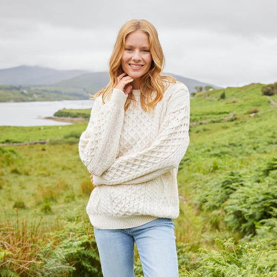 Woman wearing a cream-colored fisherman-style crew neck heritage sweater with cable knit pattern, standing in a scenic outdoor landscape – lightweight and cozy knitwear for all seasons.