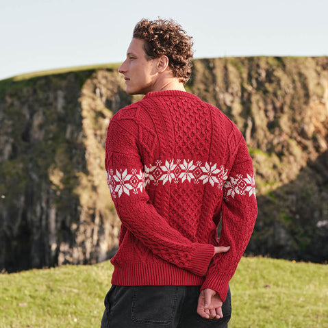 Back view of the Men's Crew Neck Aran Christmas Sweater in red, crafted with authentic Irish Aran cable knit textures and a festive white snowflake Fair Isle pattern, showcasing traditional Irish craftsmanship and classic holiday style in a scenic coastal setting.