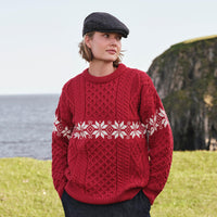 Image featuring the Women's Crew Neck Aran Christmas Sweater in red, crafted with authentic Irish Aran cable knit textures and a festive white Fair Isle snowflake pattern, blending traditional Irish craftsmanship with timeless holiday style along a rugged coastal setting.