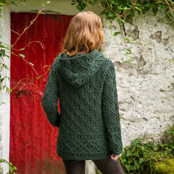 A person with shoulder-length wavy red hair is seen from behind, wearing a dark green aran hoodie. They stand in front of a weathered white stone wall, partially covered in greenery, with a vibrant red door visible on the left side of the image.