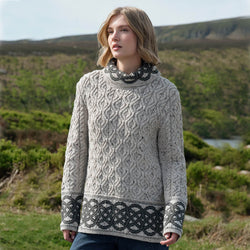 Woman wearing a grey patterned Aran sweater standing in a scenic outdoor setting with hills and water.