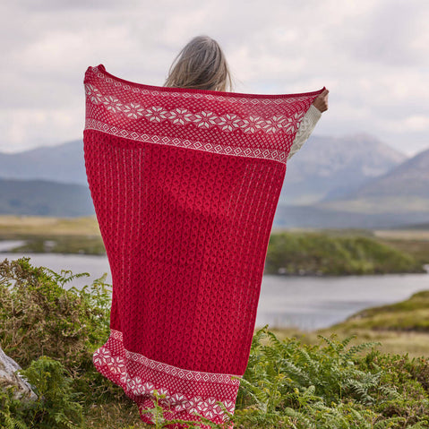 Red Aran Christmas throw with white snowflake and geometric patterns held up outdoors against a scenic lake and mountain backdrop.