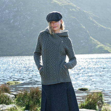 Woman wearing a gray knitted jacket and cap standing by a lake with mountains in the background