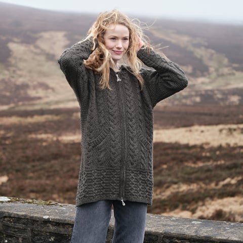 Woman wearing a long charcoal grey Aran wool cardigan with a hood and full zip, featuring cable and honeycomb knit patterns, set against a scenic Irish mountain landscape