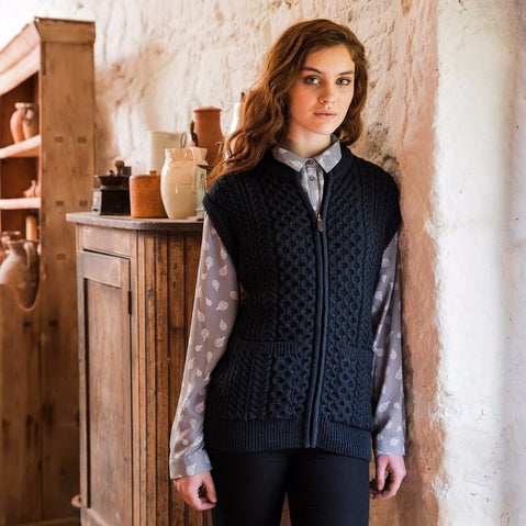 A woman standing in a room with wooden furniture wearing a traditional sleeveless zipper vest with Aran stitch work in gray.
